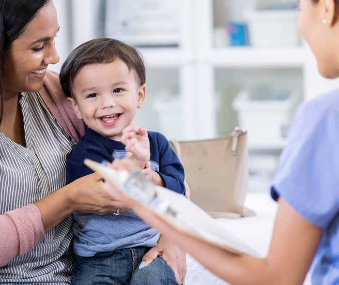 Kleiner Junge sitzt auf dem Schoß seiner Mutter gegenüber von einer Arzthelferin und freut sich  Kleiner Junge sitzt auf dem Schoß seiner Mutter gegenüber von einer Arzthelferin und freut sich